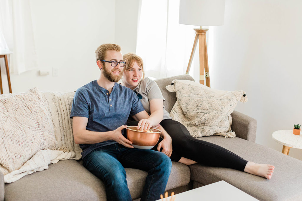 14 At Home Date Ideas - movie marathon. Sam and Jeff curled up on a grey couch eating popcorn together